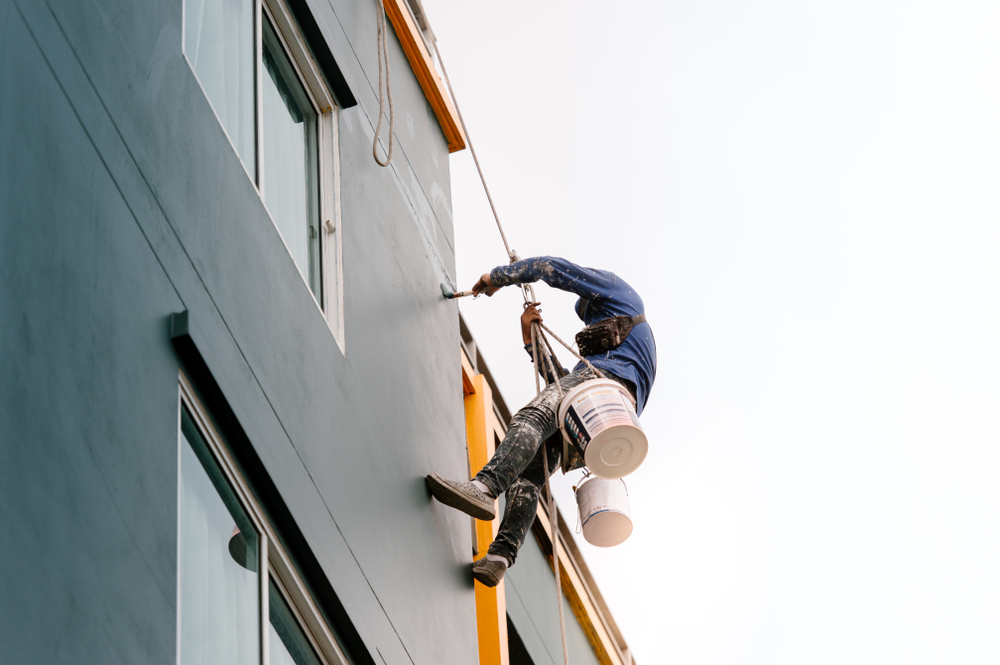 A painter uses a rope system to suspend themselves in the air to paint the exterior of a commercial property. There is a white sky in the background.