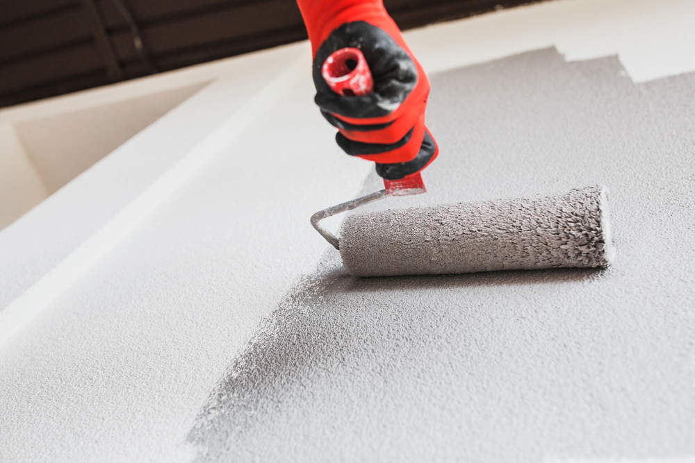 A person wearing a black and red glove uses a paint roller to paint a white wall gray.